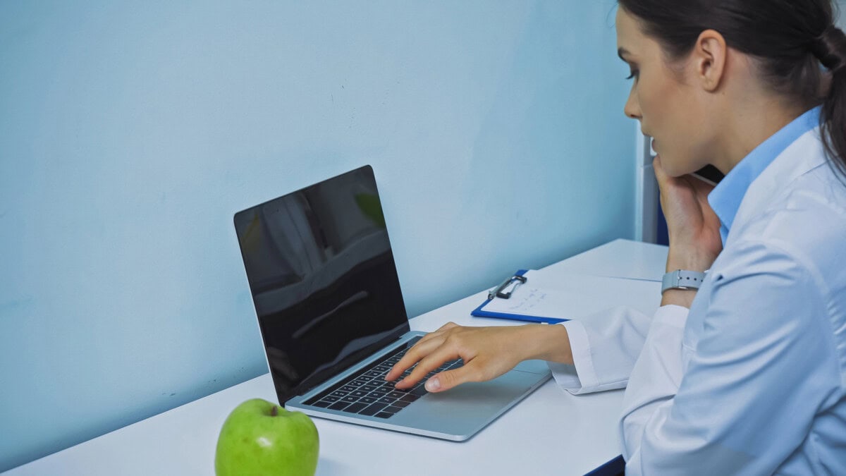 Stomatologist typing on laptop and talking on smartphone near fresh apple and clipboard